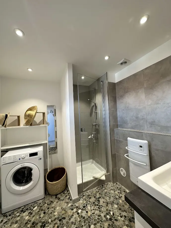 Salle de bain moderne avec douche en verre, carrelage gris, sol en galets et lave-linge à Loc'Amargue aux Saintes-Maries-de-la-Mer.