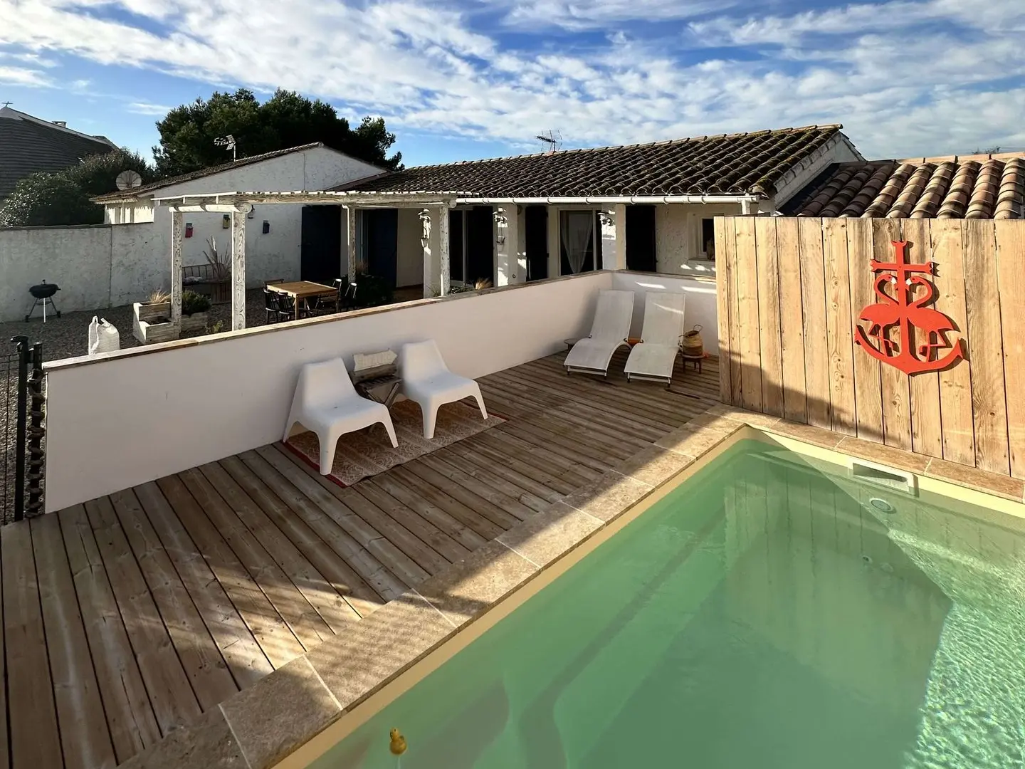 Une arrière-cour accueillante avec une terrasse en bois, une piscine et des meubles d’extérieur modernes, sous un ciel bleu clair.