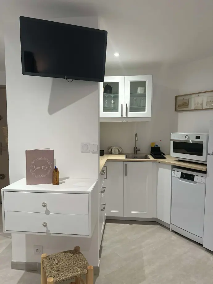 La cuisine de l'appartement « La Rose des Vents » offre un espace moderne et fonctionnel, parfaitement aménagé avec des équipements complets : lave-vaisselle, micro-ondes, réfrigérateur et de nombreux rangements. Une télévision murale est également installée pour profiter de vos émissions préférées pendant vos repas. Cuisine moderne et équipée d'un appartement, avec lave-vaisselle, micro-ondes, évier et meubles blancs, accompagnée d'une télévision murale
